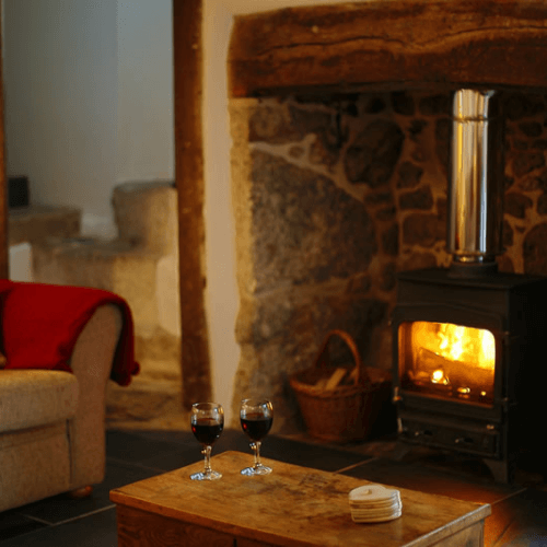 Cosy lounge with glasses of wine in front of a roaring fire set within the inglenook fireplace of Three Pound Cottage the Dartmoor thatched holiday cottage