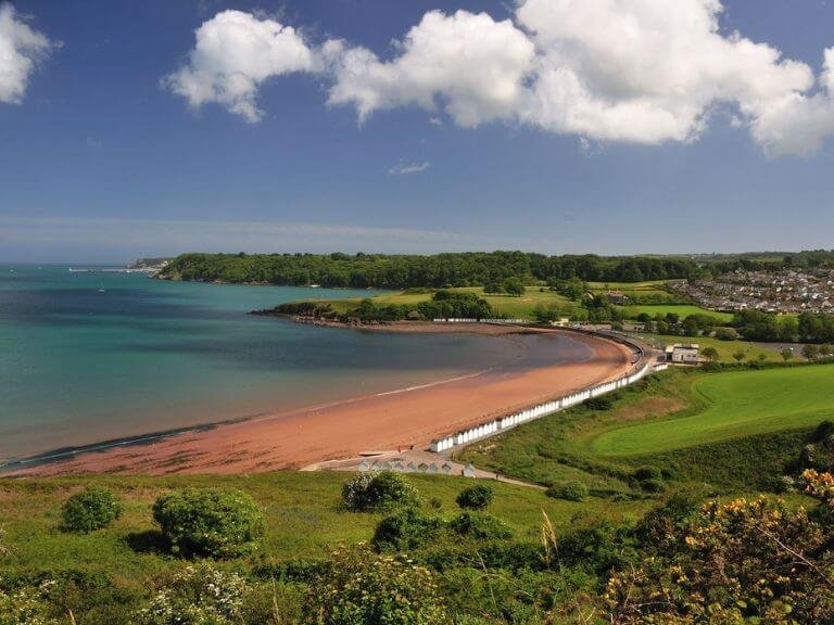 long sandy beach with calm pale blue sea bordered by lush green countryside